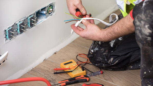 person wearing a work uniform holding electrical wires near wall outlets surrounded by various tools for electrical work with focus on two wire connections