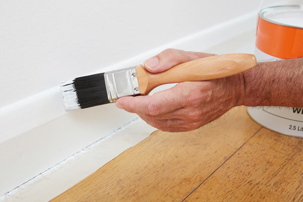 Person using a paintbrush to paint the edges of a wall, next to a can of paint on a wooden floor. Ideal for home improvement projects.